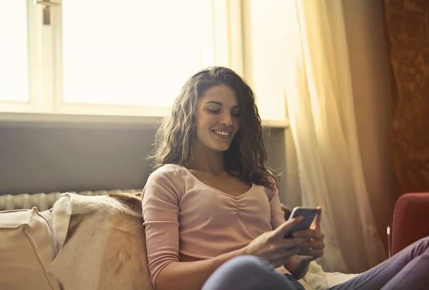 בחורה צעירה יושבת עם הגב לחלון וקוראת דברים בתוך הסמארטפון שלה עם חיוך