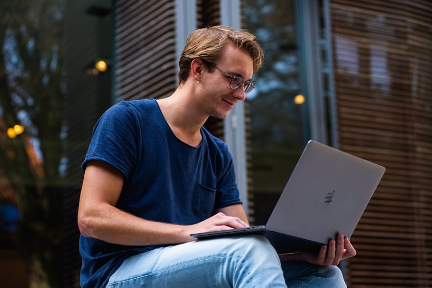 בחור צעיר ובלונדיני עם משקפיים יושב בחוץ ובודק דברים במחשב שלו עם חיוך