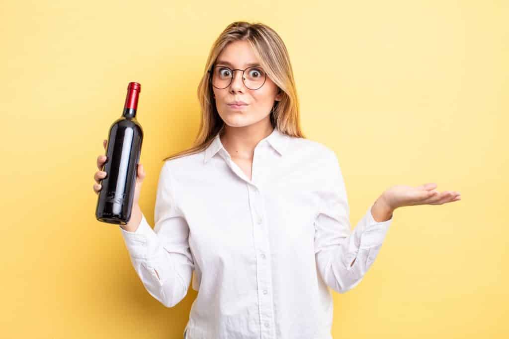 בחורה בלונדינית עם משקפיים בחולצה לבנה מחזיקה בקבוק יין ומושכת בכתפיים