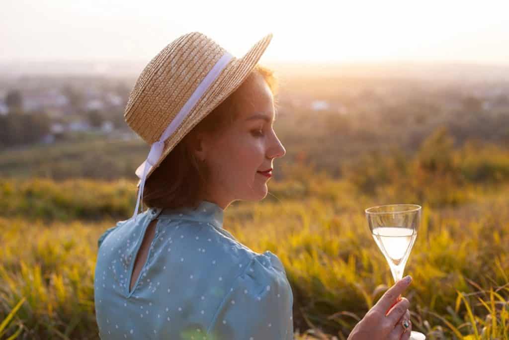 בחורה עם כובע קש יושבת בטבע מול שקיעה ומחזיקה כוס יין לבן