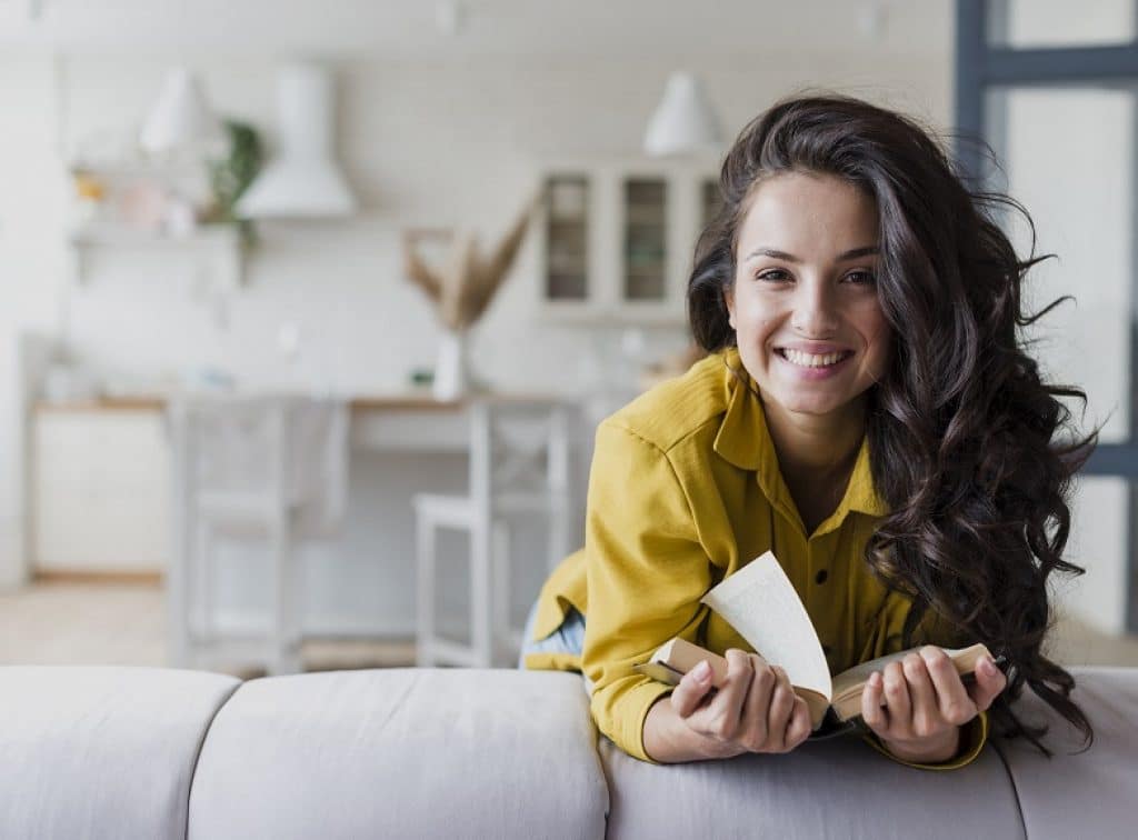 בחורה עם שיער שחור וחולצה צהובה מחייכת מחזיקה ספר ונשענת על ספה