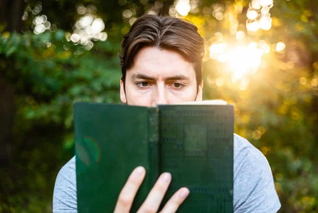 בחור מחזיק ספר בצבע ירוק וקורא עם מבט מתוח ברקע עצים ושמש