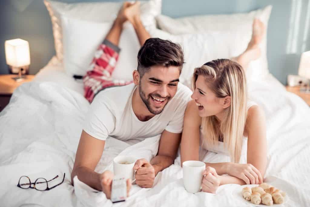 גבר ואישה מחייכים שוכבים על הבטן במיטה לבנה בחופשה