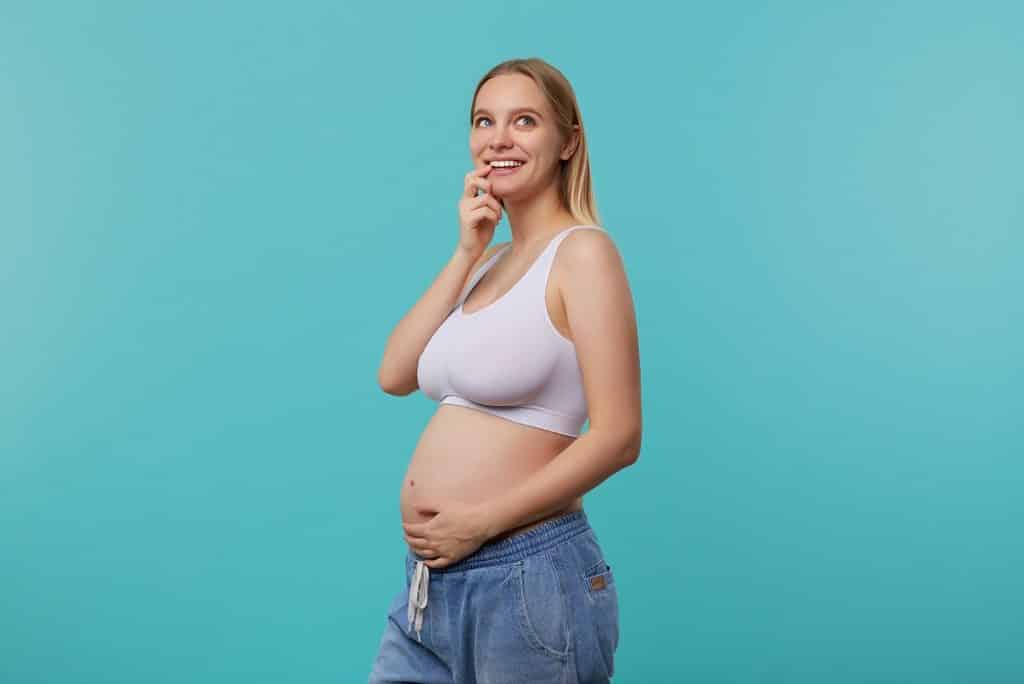 אישה בהריון מחייכת וחושבת יד על הסנטר רקע תכלת