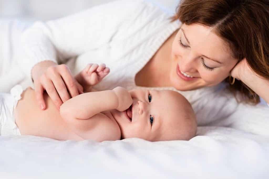 אישה עם חולצה לבנה ותינוק עם יד בפה מחייכים ושוכבים על מיטה