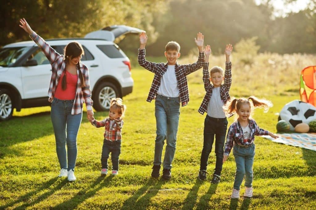 אמא וארבעה ילדים עומדים בדשא ומחייכים ברקע רכב ומשחקים