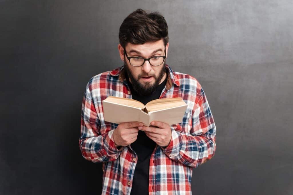 בחור במשקפיים וחולצה משובצת מחזיק ספר וקורא מרותק רקע אפור