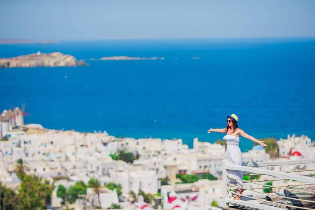 אישה בשמלה לבנה עומדת במרפסת שמשקיפה על הנוף של יוון