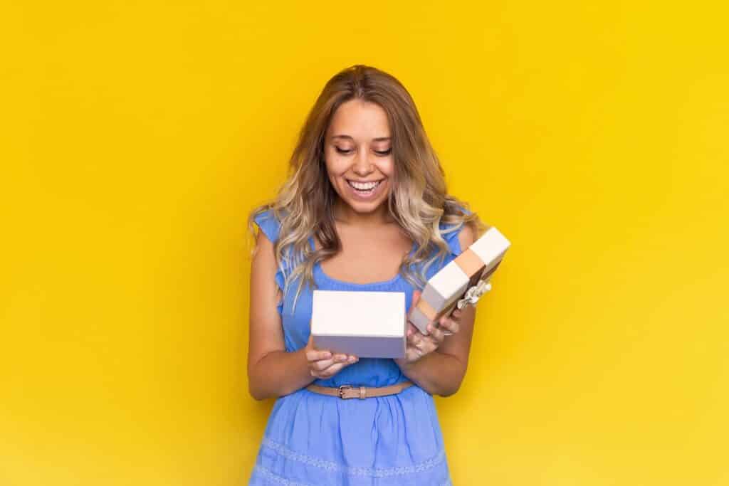 בחורה בשמלה תכלת מחזיקה קופסת מתנה ומחייכת רקע צהוב