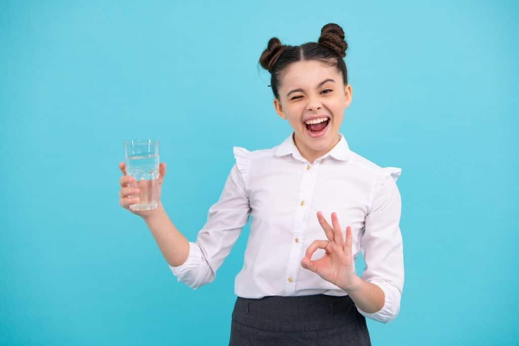 ילדה בחולצה לבנה מחזיקה כוס מים מחייכת רקע תכלת