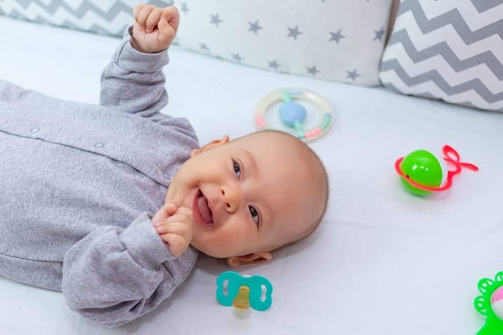 תינוק מחייךעם אוברול אפור שוכב בלול וסביבו משחקים ומוצץ