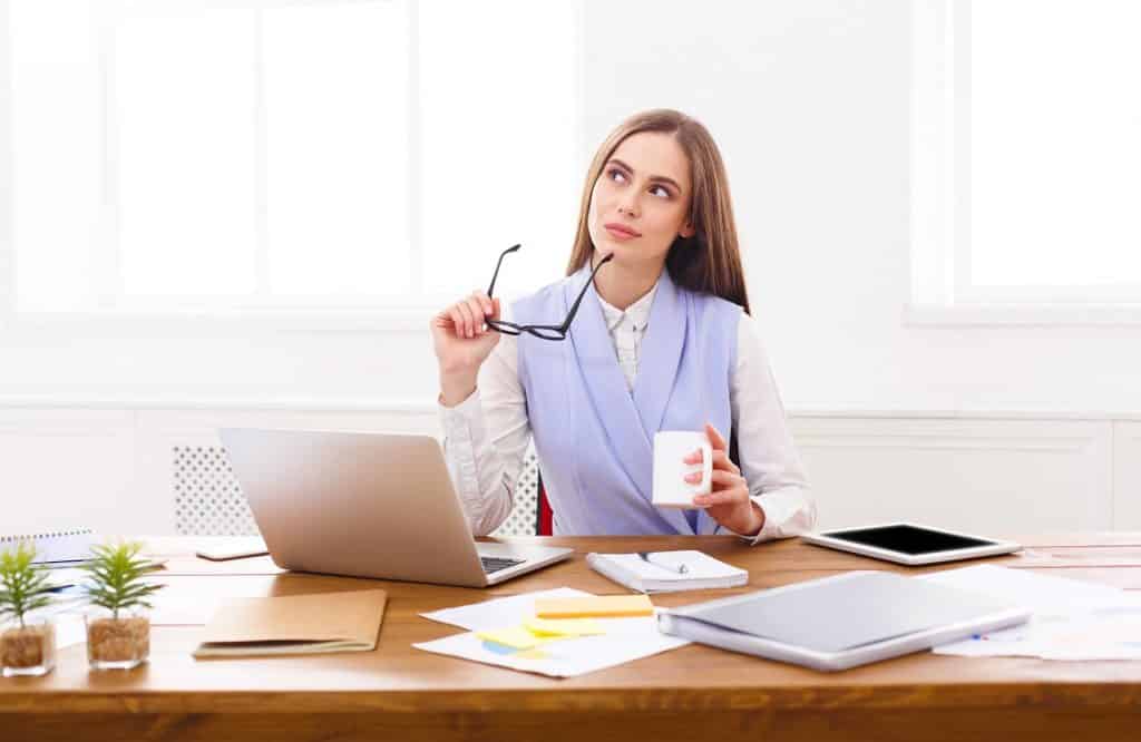 אישה עם סוודר סגול יושבת מול מחשב נייד מחזיקה משקפיים וחושבת