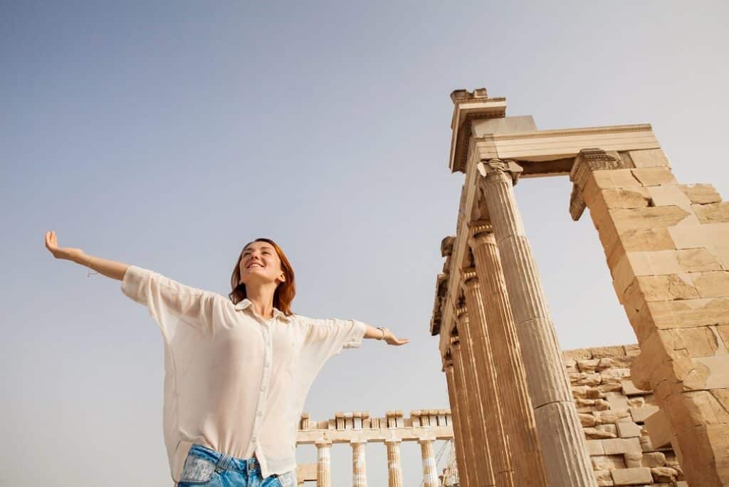 בחורה מחייכת בידיים פרושות וחולצה לבנה וברקע האקרופוליס באתונה יוון