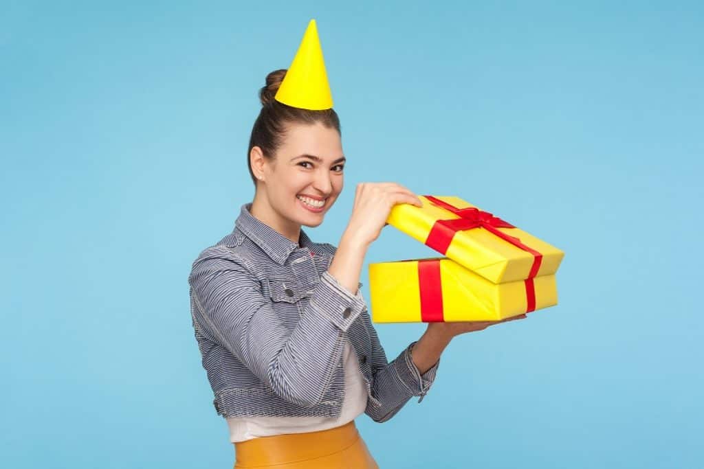 בחורה עם כובע יום הולדת מחייכת ומחזיקה מתנה צהובה עם סרט אדום רקע תכלת