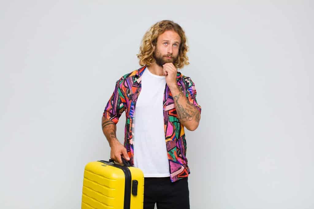 בחור בלונדיני עם שיער ארוך וחולצה צבעונית מחזיק מזוודה וחושב רקע לבן