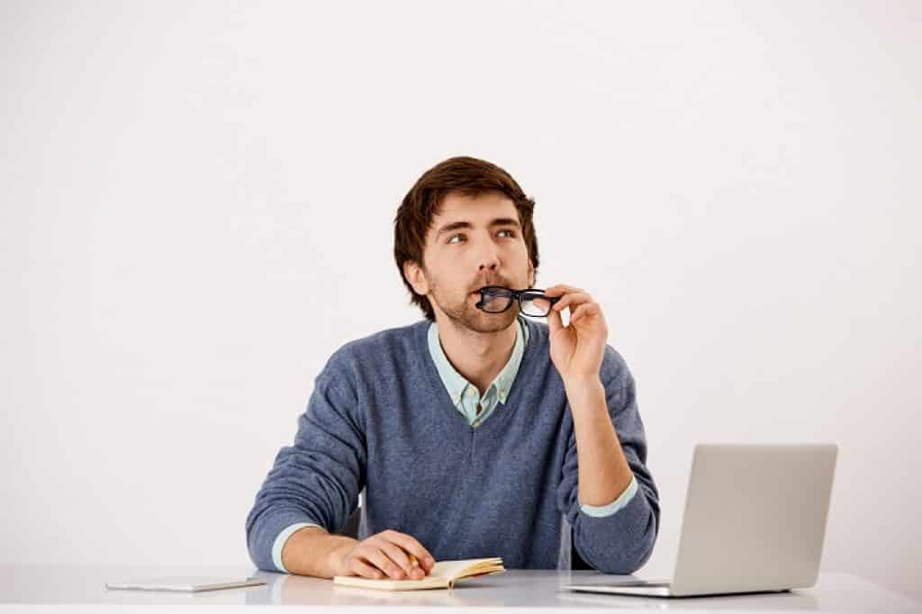 בחור בסוודר כחול יושב ליד מחשב נייד מחזיק משקפיים במבט חושב