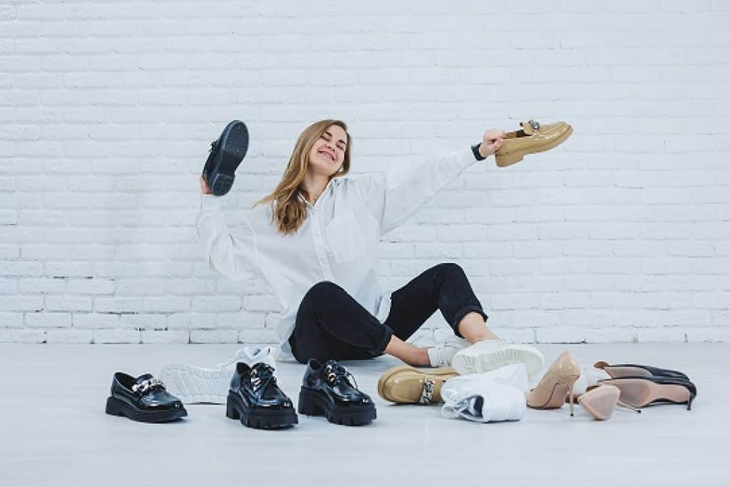 אישה מחיכת ושמחה מחזיקה נעליים יושבת על הרצפה עם נעליים סביבה רקע לבן