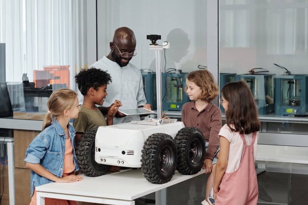 מורה ותלמידים עומדים סביב רובוט על גלגלים ברקע כיתה