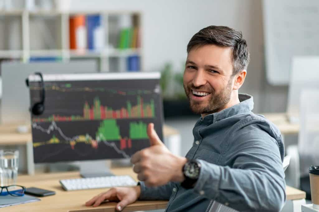 בחור יושב ליד מחשב עם מסך של מסחר בבורסה מחייך ומרים אגודל