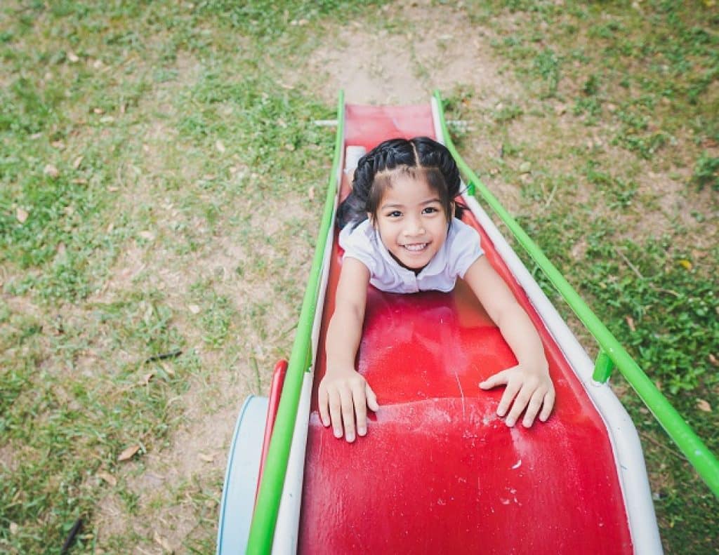 ילדה מחייכת שוכבת עם ידיים מושטות למעלה על מגלשה אדומה בדשא