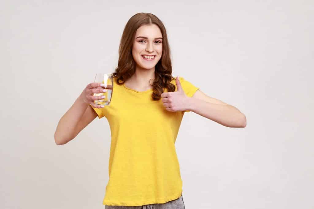 בחורה בחולצה צהובה מחזיקה כוס מים מחייכת ומרימה אגודל למעלה רקע לבן