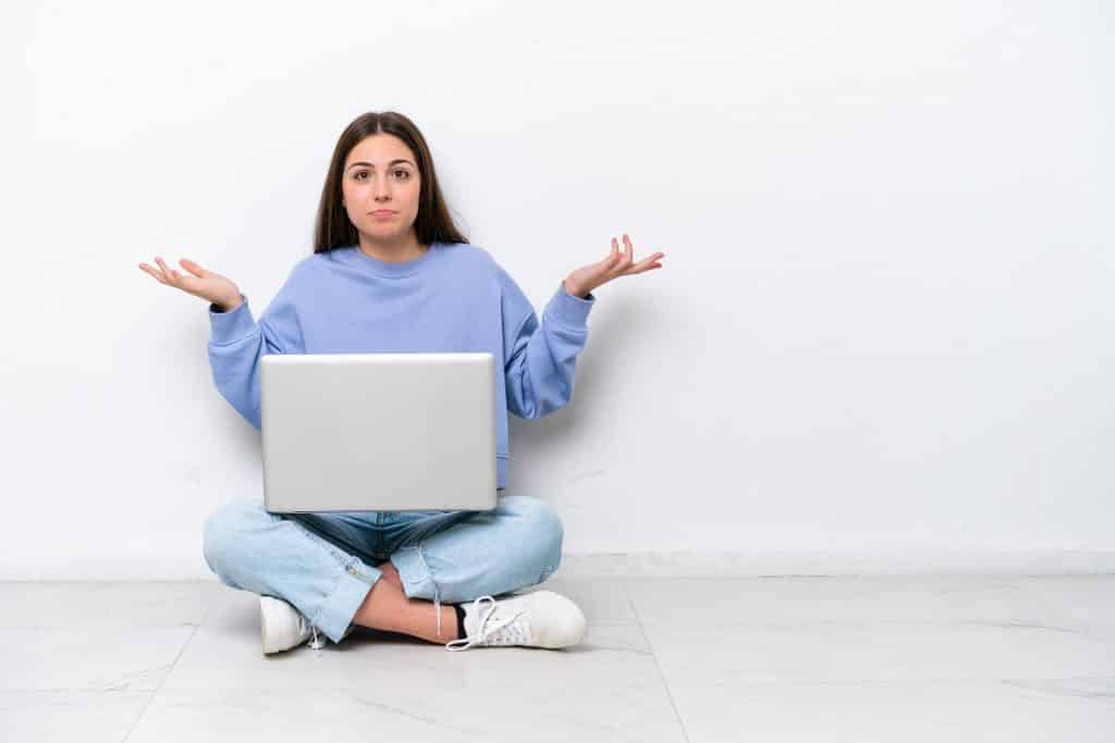 בחורה בסוודר תכלת יושבת על הרצפה עם מחשב נייד מבט מבולבל רקע לבן