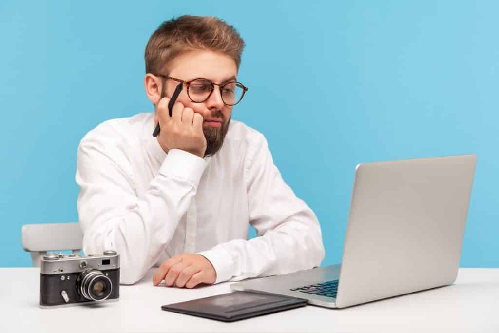 בחור בחולצה לבנה ומשקפיים מחזיק עט ציור ללוח גרפי מול מחשב מבט חושב