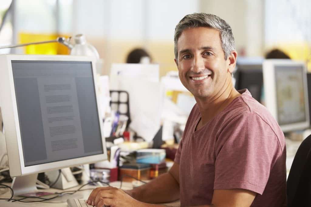 גבר בחולצה בורדו קצרה יושב ליד מחשב במשרד מחייך