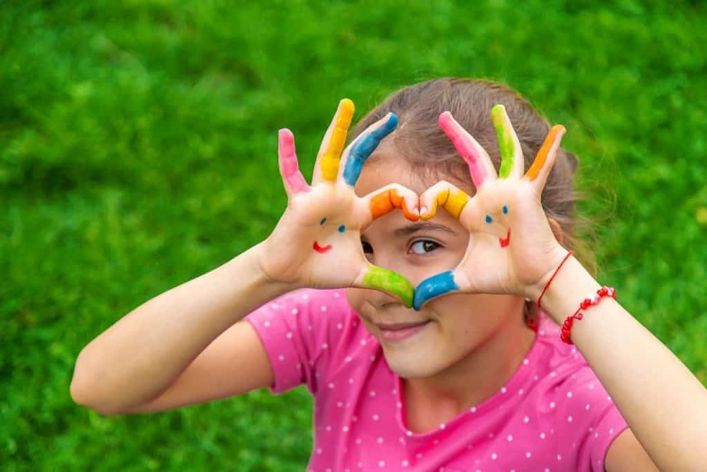 ילדה בחולצה וורודה מחזיקה את הידיים בצורת לב מעל העין אצבעות צבועות דשא