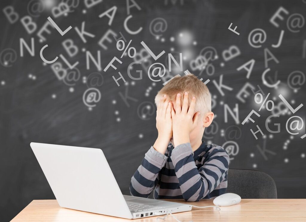 ילד בלונדיני יושב מול מחשב נייד עם ידיים על הפנים ברקע לוח עם מספרים ואותיות