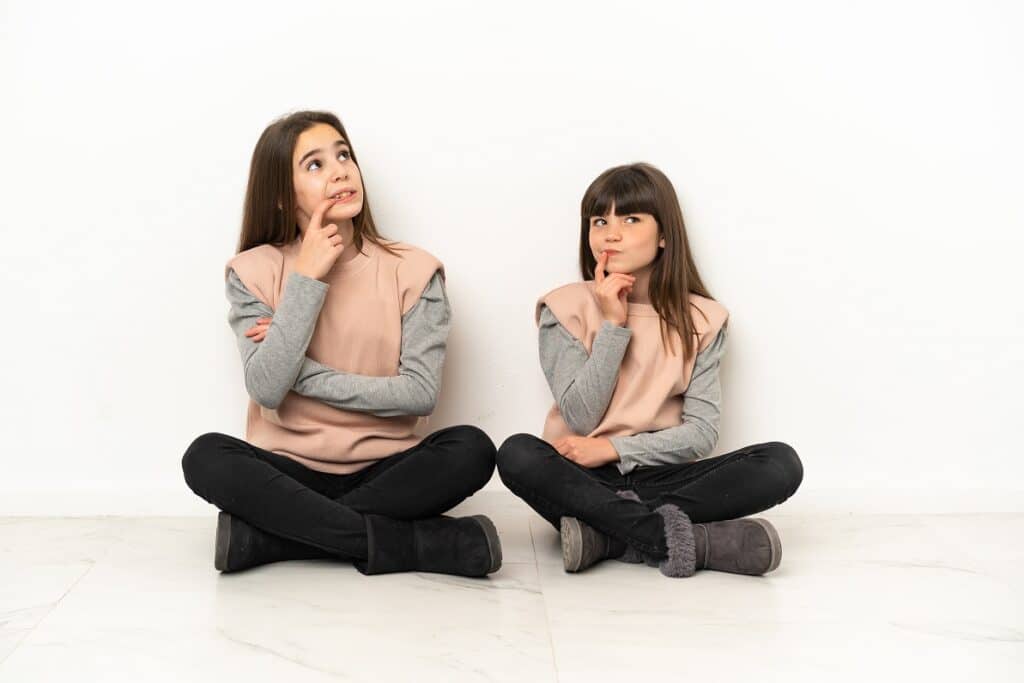 שתי אחיות לבושות בבגדים זהים יושבות על הרצפה וחושבות רקע לבן
