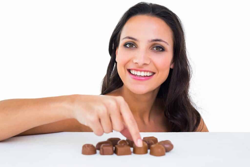 בחורה מחייכת נוגעת בפרלינים של שוקולד המונחים על שולחן רקע לבן