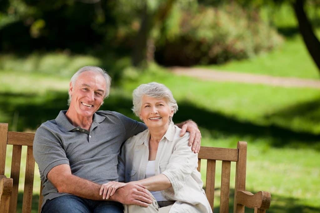 גבר ואישה מבוגרים יושבים על ספסל מחייכים ומחובקים רקע של דשא ועצים