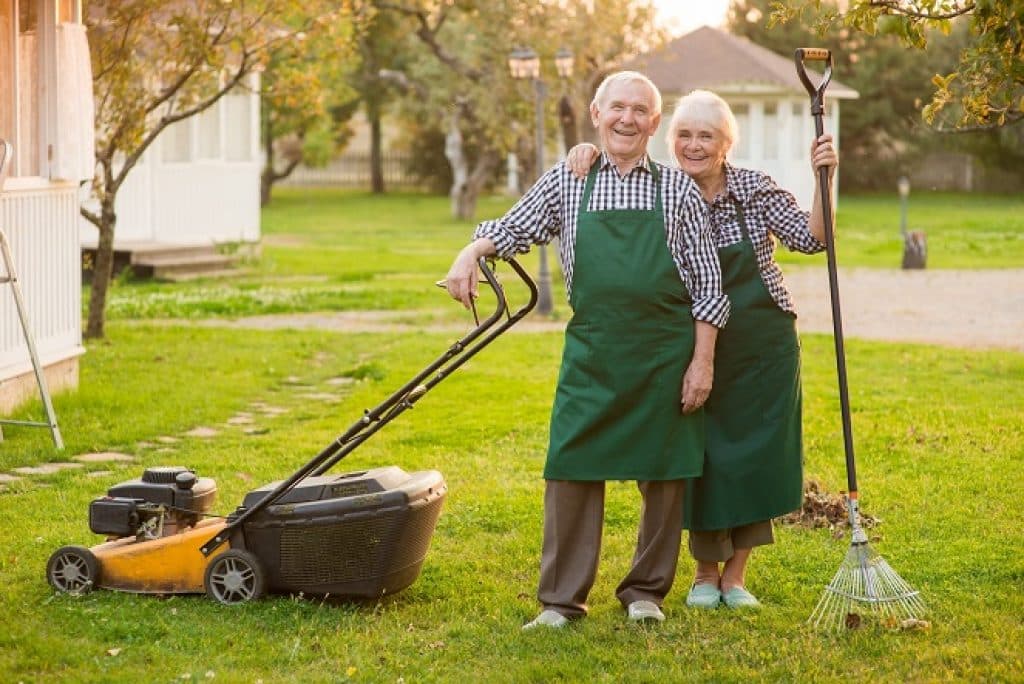 גבר ואישה מבוגרים עומדים בגינה עם מגרפה ומכסחת דשא מחייכים ומחובקים
