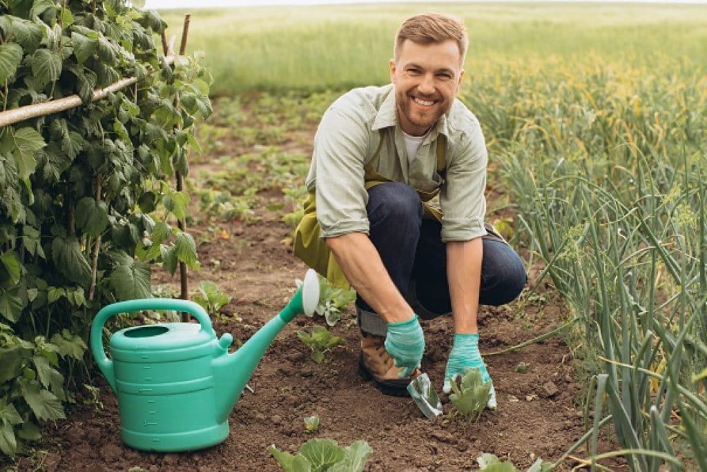 גבר מחייך עם כפפות וסינר עובד בגינה עם כף ולידו משפך ירוק