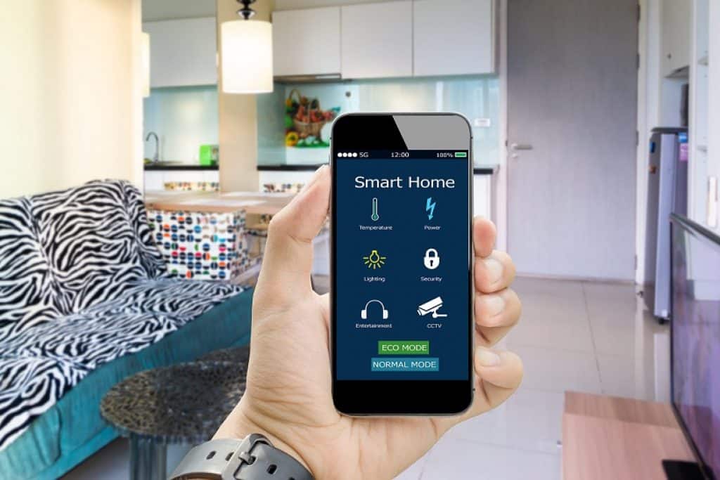 יד מחזיקה סמארטפון עם אפליקציה בית חכם סלון ומטבח ברקע