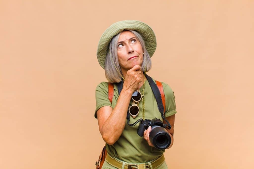 אישה עם מצלמה תיק גב משקפי שמש וכובע ירוק חושבת יד על הסנטר 