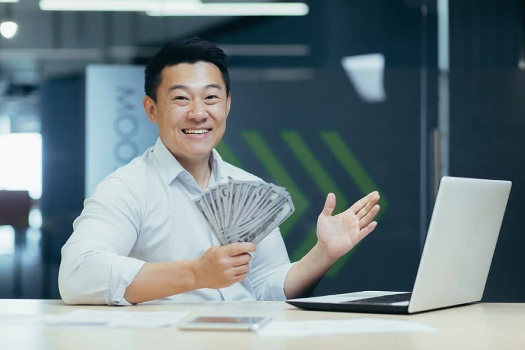 איש מחייך מחזיק חבילת שטרות יושב ליד שולחן עם מחשב נייד במשרד