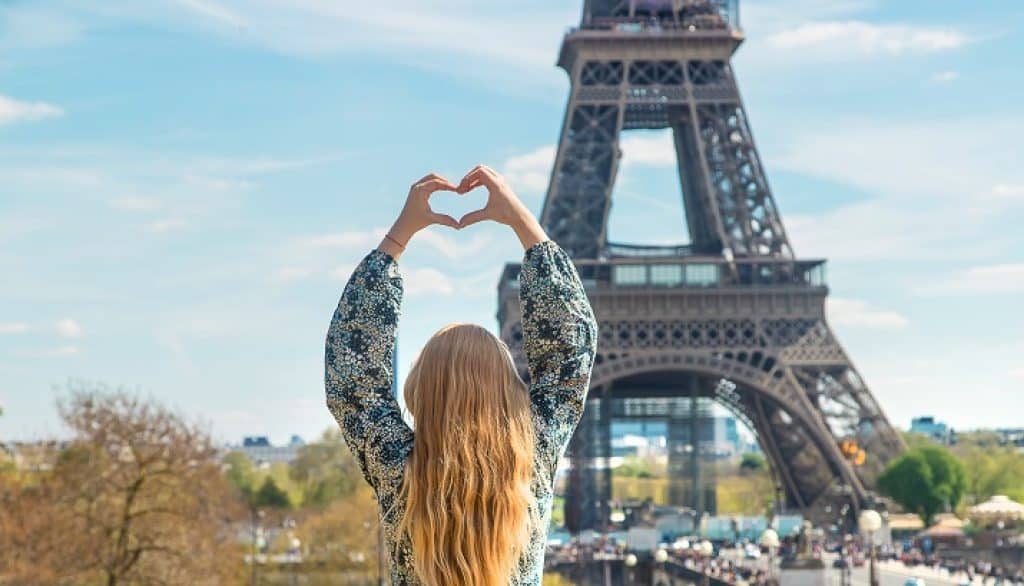 בחורה בלונדינית מחזיקה ידיים מעל הראש בצורת לב ברקע מגדל אייפל בפריז