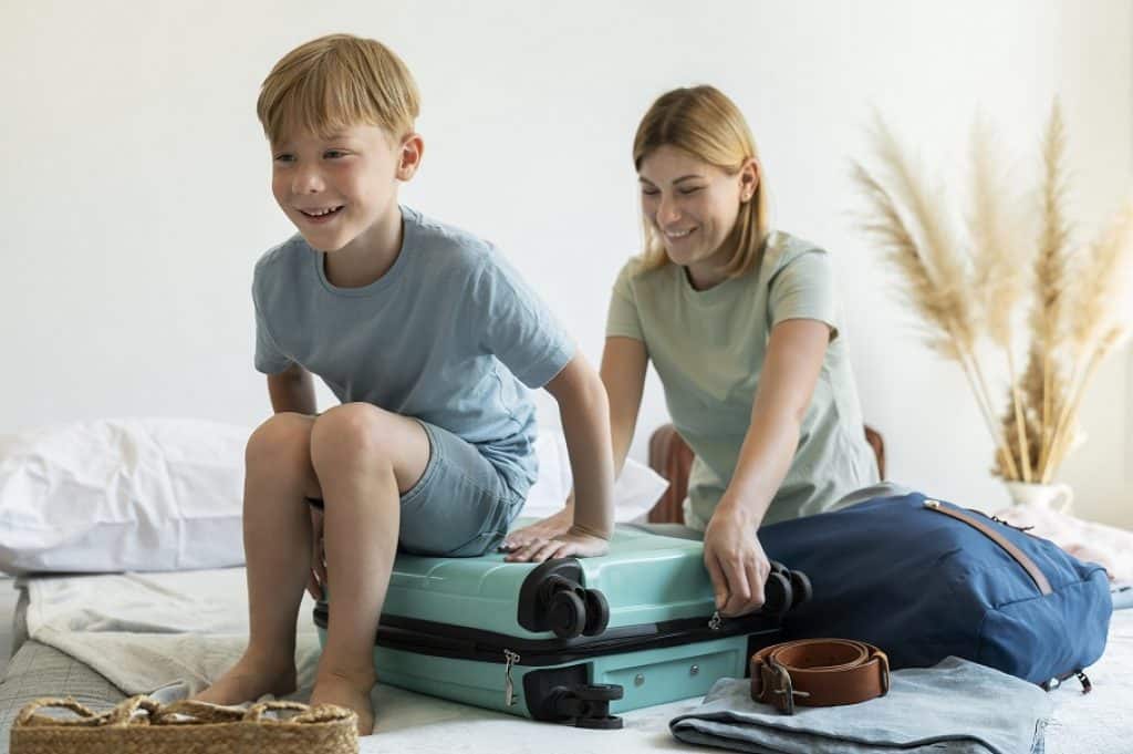 ילד מחייך יושב על מזוודה בצבע תכלת ואמא סוגרת את הרוכסן טיול בדובאי