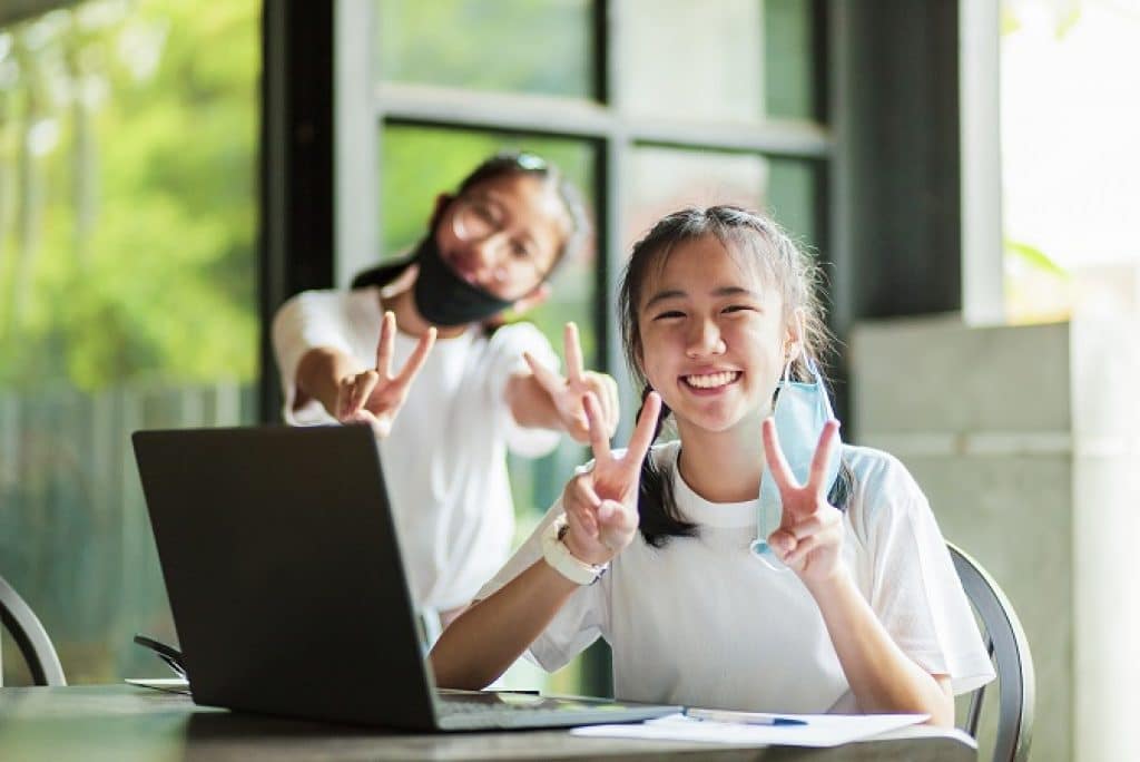נערות צעירות יושבות ליד מחשב נייד מחייכות ומרימות ידיים בסימן V