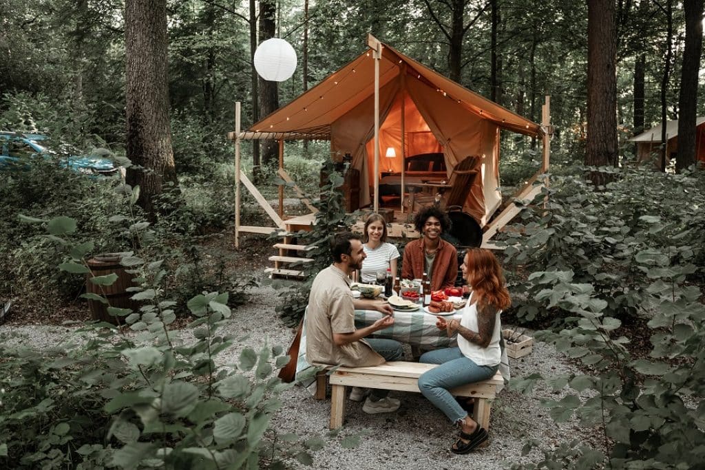 ארבעה אנשים יושבים ליד שולחן פיקניק מחייכים ברקע אוהל גלמפינג בטבע