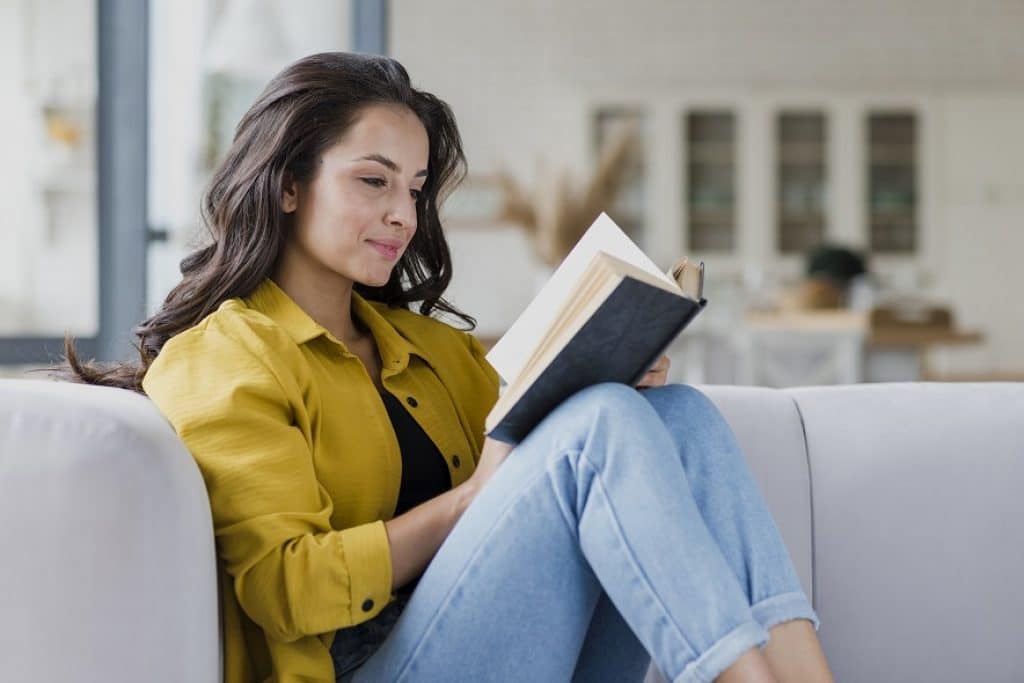 בחורה בחולצה צהובה מחייכת ויושבת על ספה קוראת ספר