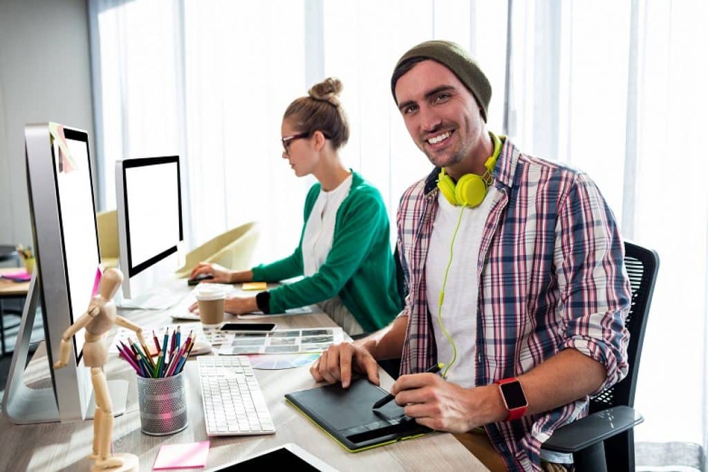 בחור מחייך עם אוזניות צהובות וחולצה מכופתרת יושב מול מחשב