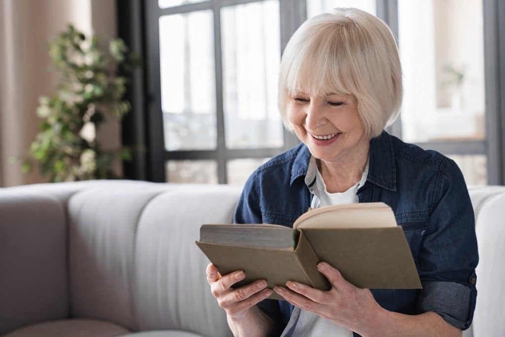 אישה מבוגרת יושבת על הספה קוראת ספר דרמה ומחייכת