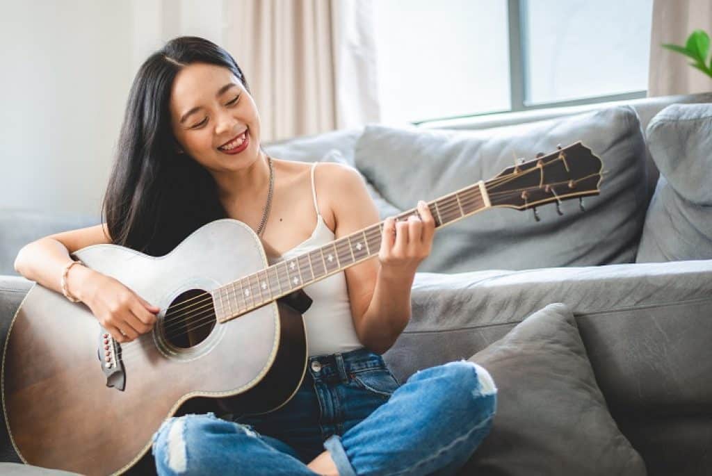 בחורה אסייתית מחייכת יושבת ליד ספה מנגנת בגיטרה אקוסטית