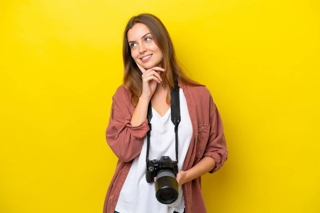 בחורה מחזיקה מצלמה מחייכת עם יד על הסנטר חושבת רקע צהוב
