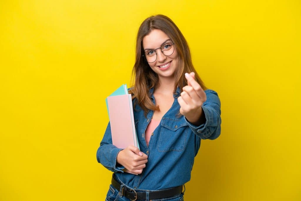 בחורה עם משקפיים מחייכת מחזיקה מחברות ומושיטה יד קדימה רקע צהוב