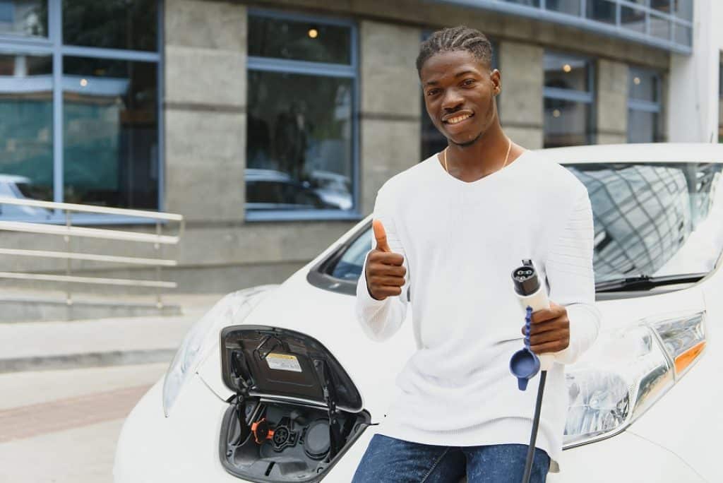 בחור אפריקאי מחייך עומד ליד רכב חשמלי עם פתח טעינה פתוח