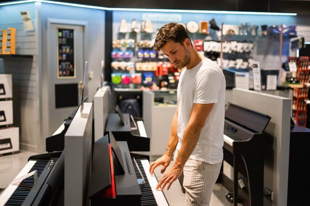 בחור בחולצה לבנה עומד ליד פסנתר חשמלי בחנות מוזיקה ברקע פסנתרים